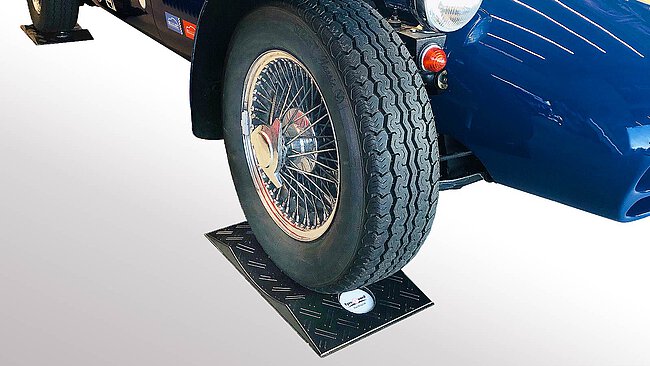 a black convex-shaped TyreGuard® brand tyre protection made of high-strength plastics with non-slip protection mat, placed underneath a blue historic sports roadster