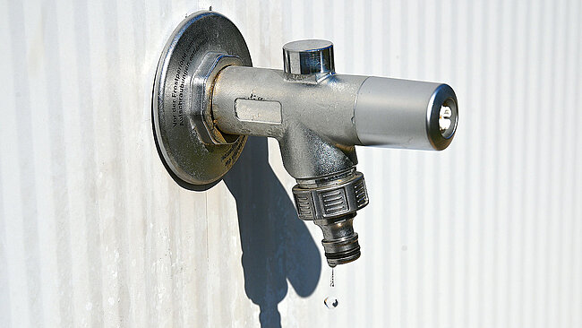 a water tap made of silvery-shining metal, fixed to a grey striped panel wall, with a drop of water dripping from the hose connector