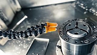 two flexible black coolant hoses of the Aqua-Loc series in plastic, with orange-coloured nozzle tips, aligned towards a turning spindle with jaw chuck inside a metal-clad chamber of a CNC machine tool