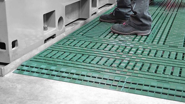 a lay-out of dark-green modular plastic floor gratings with ramp pieces, in front of a grey CNC machine tool with a machine operator standing on the floor gratings