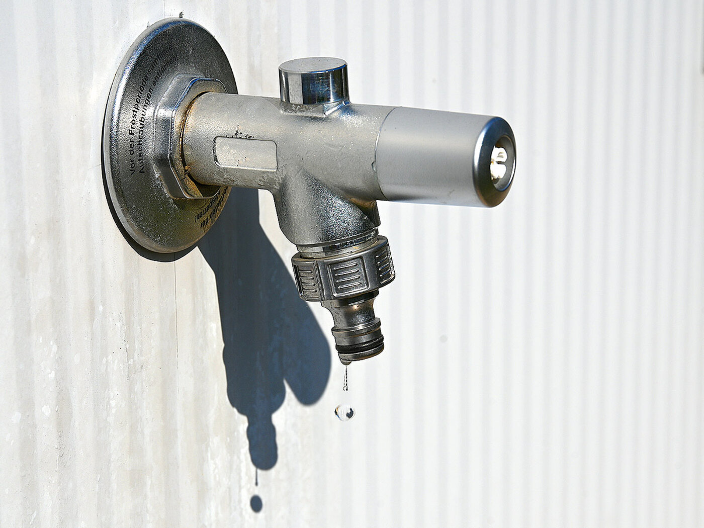 a water tap made of silvery-shining metal, fixed to a grey striped panel wall, with a drop of water dripping from the hose connector